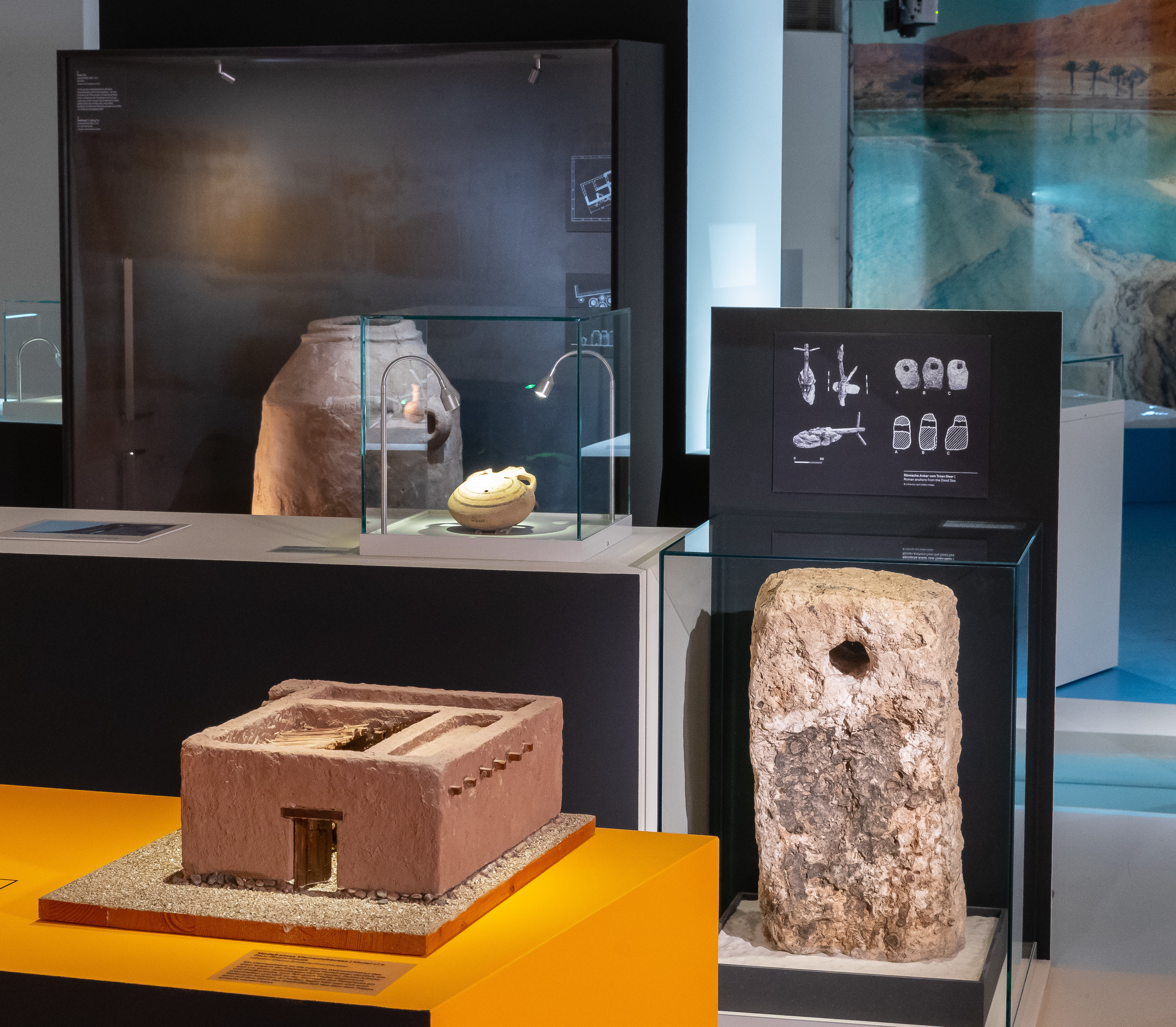 Blick in die Ausstellung, im Vordergrund das Modell eines Hauses mit Flachdach, in der Bildmitte eine Steinanker, im Hintergrund eine Vitrine mit großen Keramikgefäßen