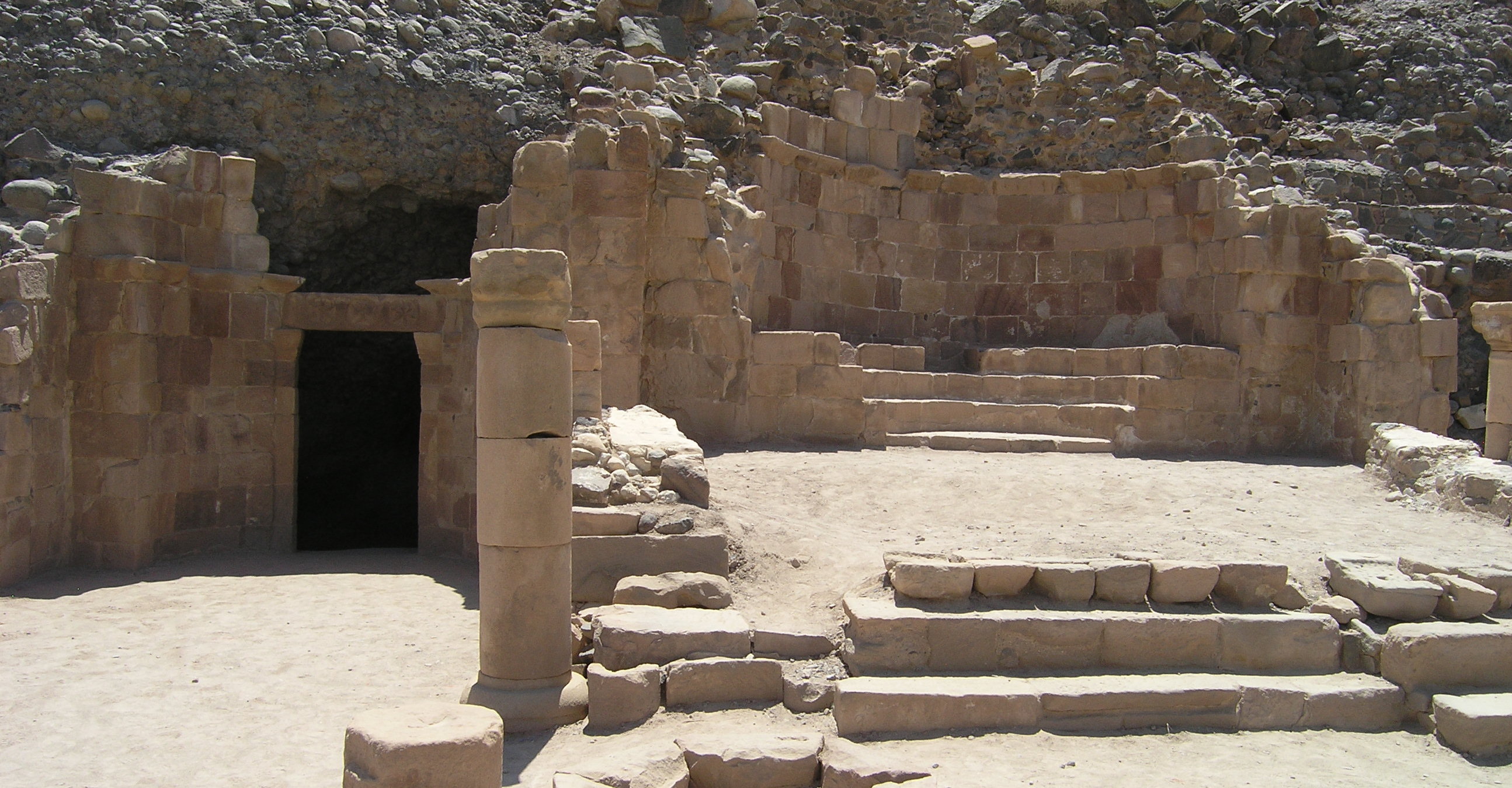 Zu sehen ist ein ruinenartiger Bereich vor einem Felsen. In der rechten Bildhälfte sind Grundrisse eines Gebäudes mit Treppen darin erkennbar. Auf der linken Seite führt ein dunkler Eingang in den Berg hinein.